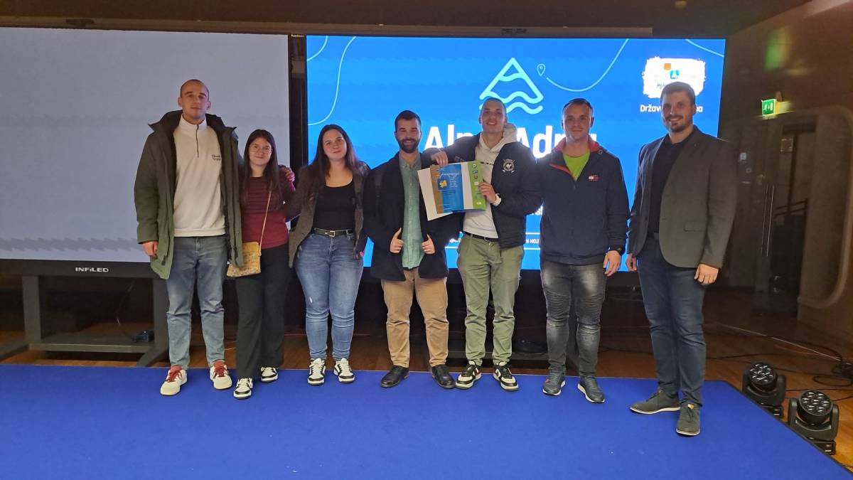 Sedem zadovoljnih študentov in študentk stoji pred panojem. V rokah držijo priznanja.