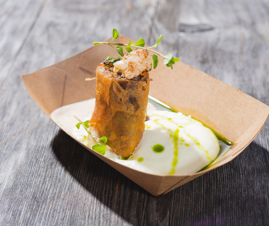 Gurmanska jed na leseni mizi vključuje hrustljavo žemljico, obloženo z zelišči in kozicami, servirano na kartonskem pladnju s kremasto omako in zelenimi oljnimi poudarki.