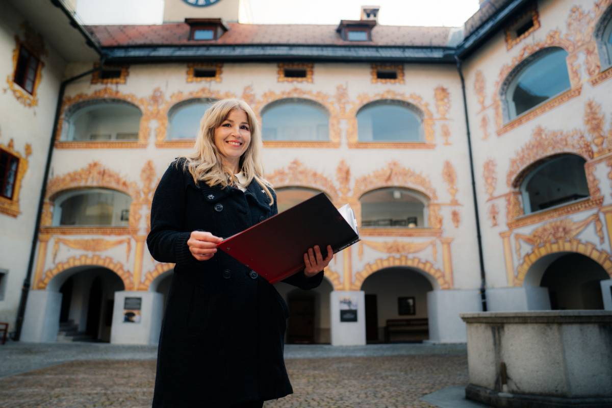 Urejena gospa srednjih let z mapo v roki stoji na grajskem dvorišču.
